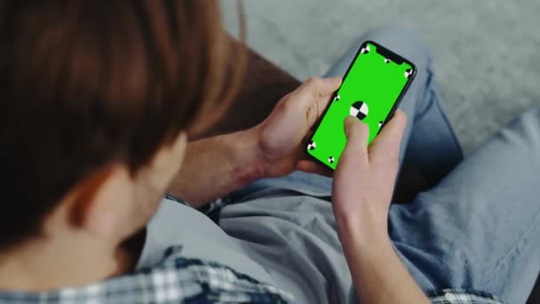 白人男子坐在客厅里 手里拿着装有绿色彩色按键模拟屏幕的智能手机 刷着显示屏 上网社交媒体 网上购物 — 图库视频影像