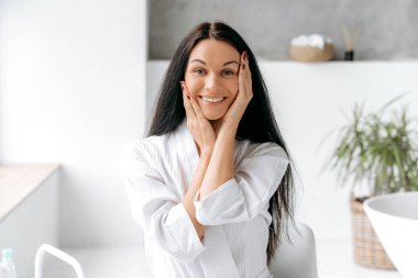 Uzun siyah saçlı, güzel, bakımlı beyaz bir kadının fotoğrafı. Banyoda duruyor, ellerini yüzüne yaklaştırıyor, kameraya bakıyor, mutlu bir şekilde gülümsüyor.