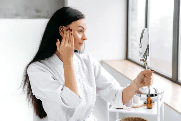 Using face cream. Happy caucasian woman, in bathrobe, holds a mirror in her hand, applies a moisturizing, anti-aging cream on her face, takes care of her skin and youth, prevents wrinkles, smile
