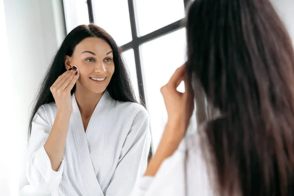 Facial cleansing. Gorgeous caucasian brunette long haired woman in white bathrobe stands in bathroom in front of a mirror, uses a white cotton pad to cleanse her sensitive skin, takes care of the skin