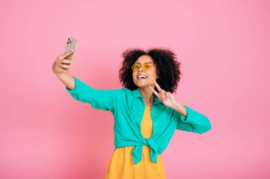 Akıllı telefondaki selfie fotoğrafı. Moda Brezilyalı kadın, yeşil günlük gömlekli, selfie çeken, parmaklarıyla barış hareketi gösteren, kamera ekranına bakan, gülümseyen, pembe arka planda duran