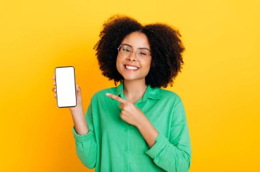 Gözlüklü mutlu Afro-Amerikan ya da Brezilyalı kadın, sunum ya da reklam için beyaz ekranlı akıllı telefonu tutuyor, ona işaret ediyor, kameraya gülümsüyor, izole edilmiş sarı arka plan