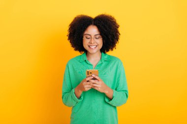 Pozitif Afro-Amerikan ya da Brezilyalı kıvırcık kadın, yeşil gömlekli, akıllı telefonunu kullanıyor, sosyal medyada arkadaşlarıyla mesajlaşıyor, online sipariş veriyor, e-postaları cevaplıyor, sarı arka planda izole edilmiş.