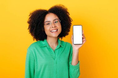 Pozitif Brezilyalı ya da İspanyol gözlüklü, rahat gömlekli, sunum ya da reklam için beyaz ekranlı akıllı telefon gösteren, kameraya gülümseyen, izole edilmiş sarı arka plan