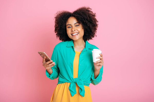 Trendy lovely stylish brazilian or hispanic woman, use her smartphone, messaging in social media, shopping online, order delivery, holds a cup of coffee, smiles at camera, isolated pink background