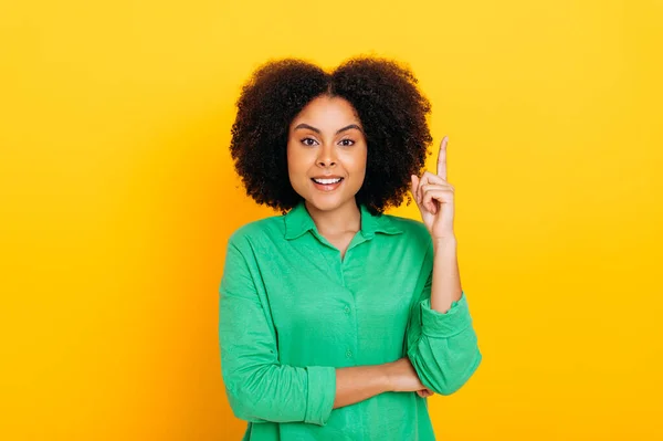 Have idea. Excited curly haired brazilian or hispanic stylish positive woman, in green shirt, looks enthusiastically at the camera, shows IDEA gesture with finger, stands on isolated yellow background