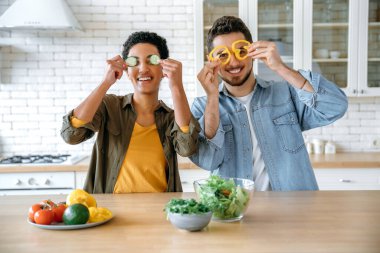 Mutlu güzel çift birlikte eğleniyor, farklı milletlerden genç adam ve kadın, mutfakta ayakta duruyor, taze sebze salatasını birlikte hazırlıyor, kamerada oynaşıyor, gülümsüyor.