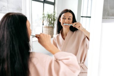 Lovely caucasian woman in a bathrobe, brushing her teeth after sleeping standing in front of a mirror in the bathroom, taking care of oral hygiene to avoid caries. Dental care, morning routine clipart