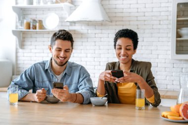 Mutlu çok ırklı çift, Afro-Amerikalı bir kadın ve beyaz bir erkek, sabah kahvaltısında cep telefonlarını evde birlikte kullanıyorlar, online video veya haber izliyorlar, mesajlaşıyorlar, gülümsüyorlar.