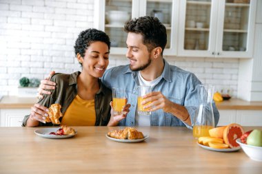 Sabah kahvaltısı. Mutlu eşler, Afro-Amerikalı bir kadın ve beyaz bir erkek, mutfakta birlikte kahvaltı ediyorlar, kruvasan yiyorlar, meyve suyu içiyorlar, iletişim kuruyorlar, birbirlerine bakıp gülümsüyorlar.