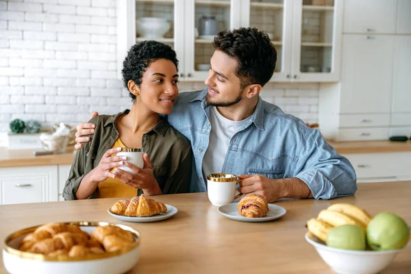 stock image Positive couple in love of different nationalities, sit und hugging at home in the kitchen, dressed in stylish clothes, drinking morning coffee with croissants, look at each other, smile