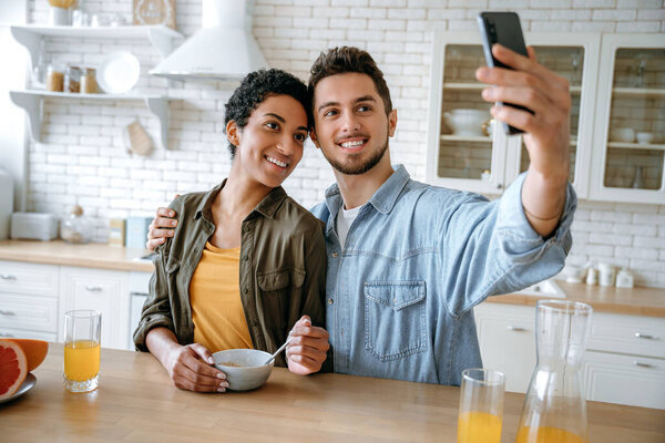 Happy beautiful couple in love, of different nationalities, bloggers, using smartphone during a morning breakfast, taking a selfie photo, looking into the camera of the mobile phone, smiling happily