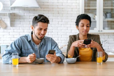 Kahvaltı konsepti. Birbirlerine aşık çok ırklı bir çift, Afro-Amerikan kadın ve beyaz erkek, sabah kahvaltısında cep telefonlarını kullanıyorlar, online video veya haber izliyorlar, mesajlaşıyorlar.