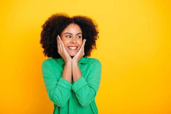 stock image Photo of a beautiful charming african american or brazilian curly haired young woman with brown eyes, wearing a green shirt, looking to the side, smiles friendly, stand on isolated yellow background