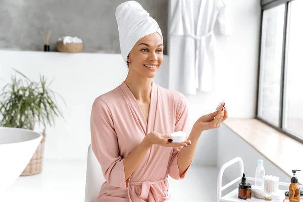 Skin cleansing. Beautiful caucasian brunette long haired woman in bathrobe with towel on head, holds a white cotton pad to cleanse her sensitive skin, uses skin care products while sit in a bathroom