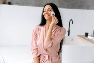 Tatmin olmuş beyaz tenli genç bir kadın, bornozlu, banyoda oturuyor, makyajdan sonra yüzünü temizliyor pamuk pedlerle, gözleri kapalı gülümsüyor. Yüz temizleme konsepti