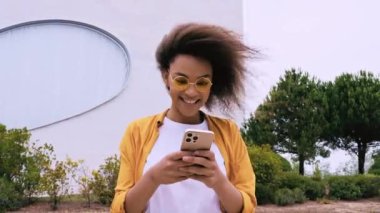 Pozitif tatmin olmuş Afro-Amerikan kıvırcık saçlı, şık giyimli, dışarıda yürürken akıllı telefonunu kullanan, sosyal ağlarda arkadaşlarıyla sohbet eden, e-postalara cevap veren, gülümseyen 