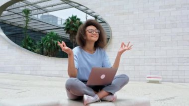 Calm relaxed curly african american young woman with glasses, office worker, sits in a lotus position outdoors near the business center, meditates, relaxes, closed her eyes. Break during online work