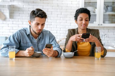 Mutlu çok ırklı çift aşık, Afrikalı Amerikalı kadın ve beyaz adam, sabah kahvaltısında cep telefonlarını kullanıyorlar, online video veya haber izliyorlar, arkadaşlarıyla mesajlaşıyorlar.
