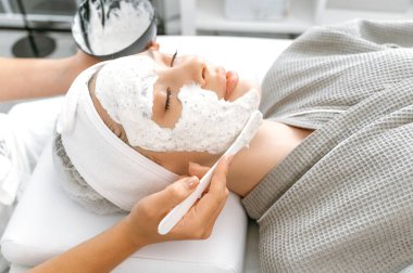 Güzellik maskeli güzel bir kadın. Güzellik uzmanı yüzüne nemlendirici maske takıyor. Nemlendirici yüz maskesi, fırça, besleyici deri. Güzellik prosedürleri, spa günü, cilt bakımı.