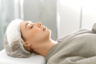 Kafkasyalı güzel bir kadının, spa salonundaki kanepede gözleri kapalı uzanmış yüz bakımı, masaj ve nemlendirici maske için hazırlanışının yan görüntüsü. Güzellik salonu
