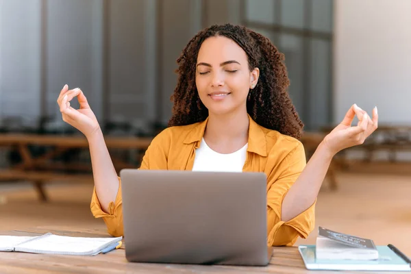 Stock image Calm relaxed brazilian or hispanic young woman with curly hair, sitting on outside with a laptop, near the university campus, meditating with closed eyes, resting from study or work online, smile