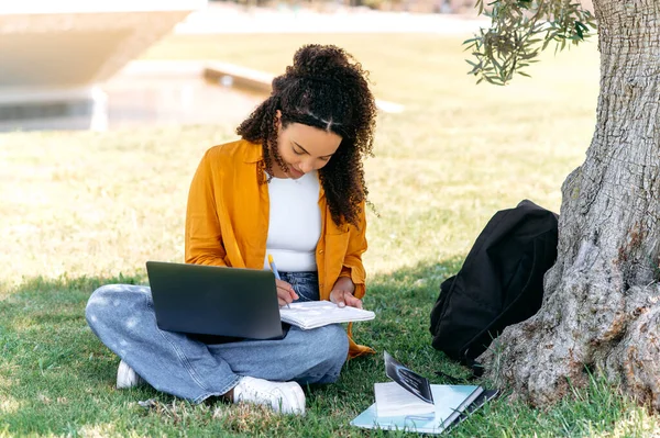 stock image Learning concept. Modern stylish positive mixed race girl student, sitting on the grass near the tree, with a laptop and notepad, doing homework, listening to an online lecture, taking notes, smile