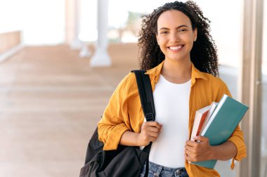 Sırt çantalı, elinde kitap ve defterlerle neşeli, kıvırcık saçlı ya da İspanyol bir kız öğrencinin yakın çekimi. Üniversite kampüsünün yanında durur ve kameraya bakar ve gülümser.