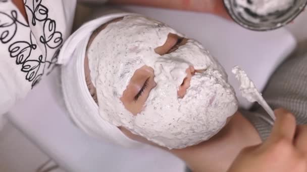 サロンで目を閉じて横になっている素敵な女性のクローズアップビデオ 彼女の顔に保湿マスクを適用する美容師 保湿栄養フェイスマスク スクラブ 栄養肌 美容方法 — ストック動画