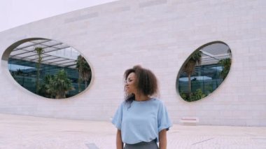 Kendine güvenen, kıvırcık saçlı, mavi gömlekli, gözlüklü, açık havada kameraya bakan ve gülümseyen Afro-Amerikan bir kadının yakın çekim videosu.