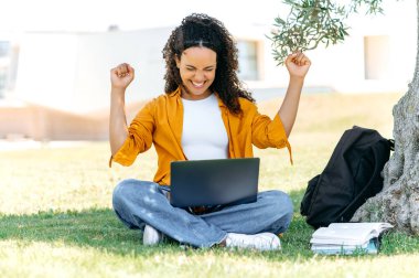 Cheerful curly mixed race female student or freelancer, sits outdoors on the grass with a laptop, rejoicing in success, passed the exam, finished the project, gesturing with both hands, smile happily