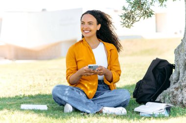 Positive stylish curly haired mixed race female student or freelancer, sits outdoors on the grass, using her smartphone, messaging in social media, texting with friends, looks away, smile happily