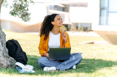 Happy smart pretty mixed race curly haired young woman, successful student or freelancer, sit outdoors on a grass with laptop, working or studying online, dreamily looks away, ponders an idea, smile