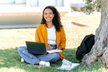 Positive smart hispanic or brazilian curly haired girl, student or freelancer, sits outdoors on a grass with a laptop, working or studying online, takes notes in her notepad, friendly smile at camera