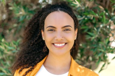 Close-up portrait of gorgeous young mixed race curly haired woman, stand outdoors, looking at camera with charming cute toothy smile, posing in casual stylish wear. Female student or freelancer