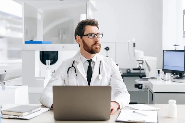 Medicine and health concept. Pensive caucasian general medicine doctor with glasses, therapist, in medical uniform, sits at workplace in hospital or laboratory, looks to the side , thinking