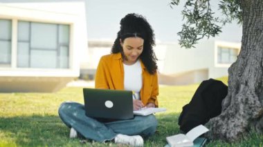 Online work or education. Positive pretty brazilian or hispanic curly girl, university student, sitting outdoors on a grass, with laptop, takes notes in a notebook, preparing for exam, smile