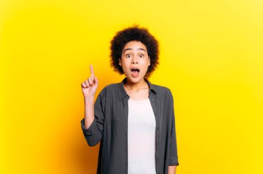 Heyecanlı Afrikalı Amerikalı ya da Brezilyalı kısa kıvırcık saçlı genç kadın, kameraya hevesle bakıyor, IDEA jestini parmağıyla gösteriyor, izole edilmiş sarı arka planda duruyor