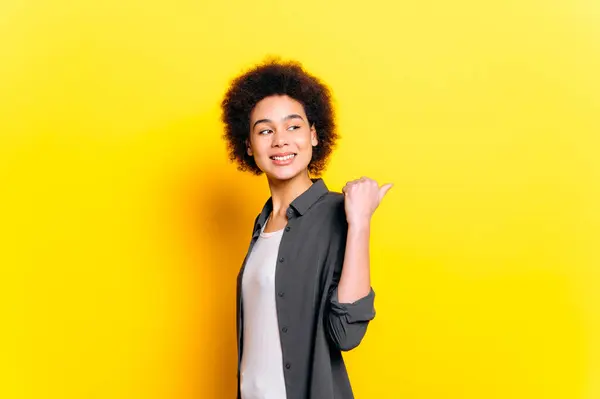 Positive african american curly haired young woman in casual wear, looks and points finger to the side behind the back, at empty copy space for presentation, stand on isolated orange background, smile