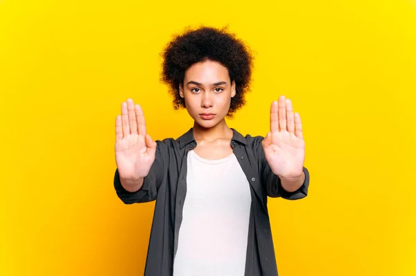 Dissatisfied frustrated african american curly haired woman, shows stop gesture with palms of two hands, against female violence, racism, abuse, stands on isolated yellow background. Stop gesture