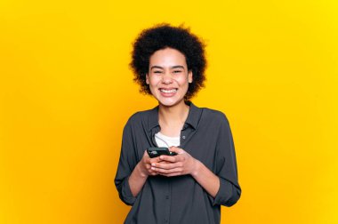 Pozitif neşeli Afro-Amerikan ya da Brezilyalı kıvırcık saçlı genç kadın, akıllı telefonunu kullanıyor, sosyal medyada arkadaşlarıyla mesajlaşıyor, çevrimiçi sipariş veriyor, e-postaları cevaplıyor, izole edilmiş sarı arka plan