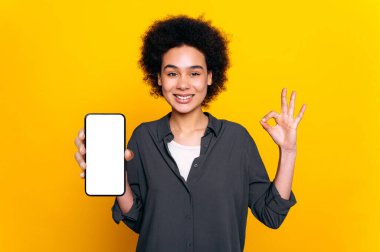 Kendine güveni tam, Afro-Amerikan kıvırcık saçlı kadın, beyaz ekranı boş akıllı telefon gösteriyor, ve el hareketleriyle kameraya bakıyor, gülümsüyor, izole edilmiş sarı arka planda duruyor.