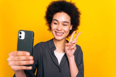 Mutlu Afro-Amerikan genç kadın, gömlek giyiyor, akıllı telefon kullanıyor, selfie çekiyor, parmaklarıyla barış hareketi gösteriyor, kamera ekranına bakıyor, gülümsüyor, izole edilmiş sarı arka planda duruyor.