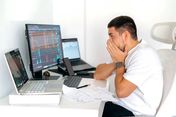 Surprised confused male crypto trader sitting at home at his work desk, shocked looks at computer screen, puzzled, had at big profits, currency growth, covering mouth with hands