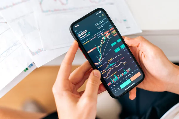 Close-up of a smartphone in male hands, with trading data index chart graph on the screen, mobile phone app analytics for cryptocurrency market. Online trading