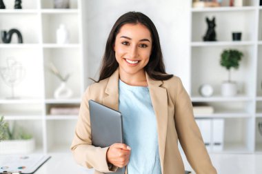 Başarılı Hintli veya Arap iş kadını portresi, sekreter, şirket yöneticisi şık bir takım elbise giymiş modern bir ofiste dizüstü bilgisayar tutuyor, kameraya bakıyor ve gülümsüyor.