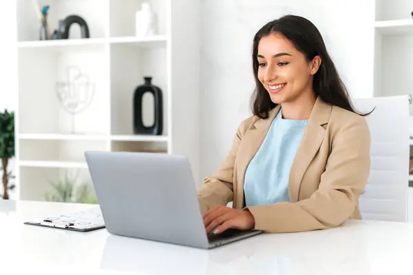 Elegant successful indian or arabian business lady, company employee sitting at workplace in the office, using laptop, working on a project, browsing and searching information in internet, smiling