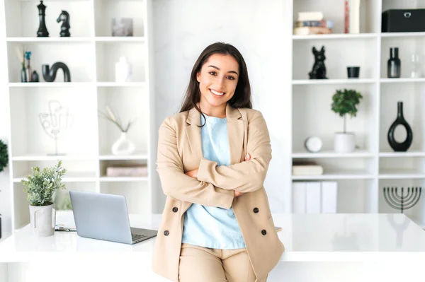 Business portrait. Amazing successful positive indian or arabian business woman, secretary, ceo manager dressed in a stylish suit standing in a modern creative office, looking and smiling at camera