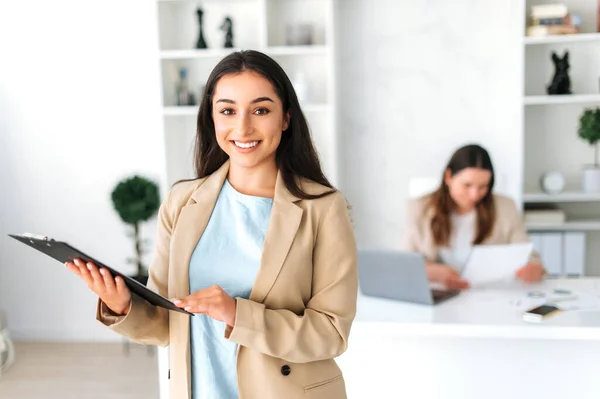 Portrait of a positive successful confident indian or arabian business woman, secretary, company executive, financial advisor standing in the modern office, holding documents, smiling friendly