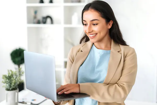 Positive successful indian or arabian business woman, secretary, manager dressed in a stylish suit standing in a modern creative office, holding an open laptop and using it, looks at screen, smiles